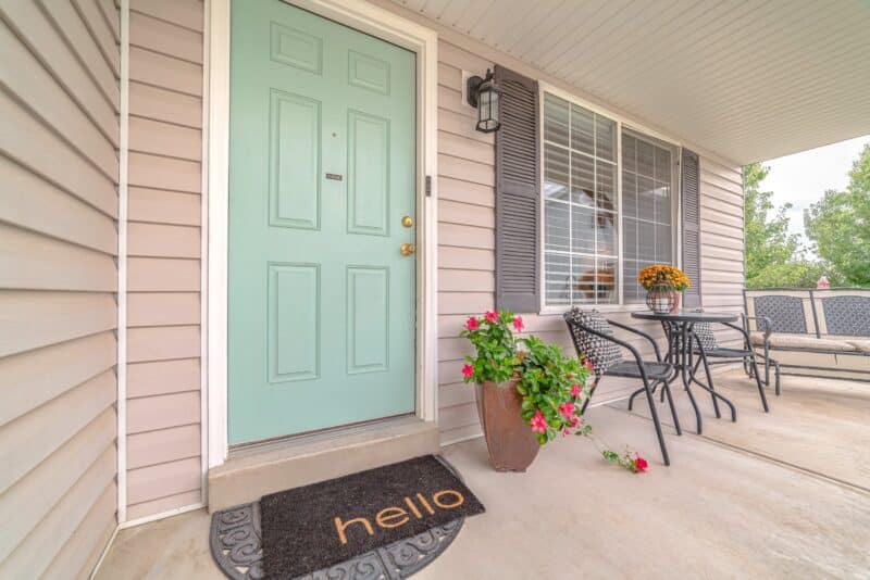 exterior of a home with a teal door and a hello door mat under it. seating areas outside right in front of the large, paned window