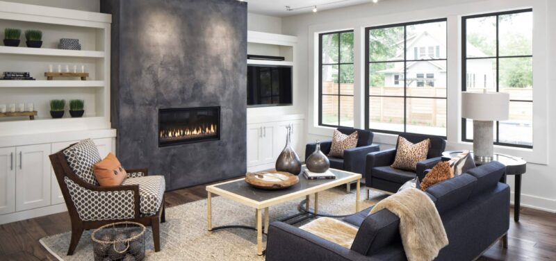 A contemporary living room with a cozy arrangement of navy sofas and a patterned armchair around a rustic wooden coffee table. Built-in white shelving, large windows providing ample natural light, and a modern fireplace with a dark surround create a warm and inviting space