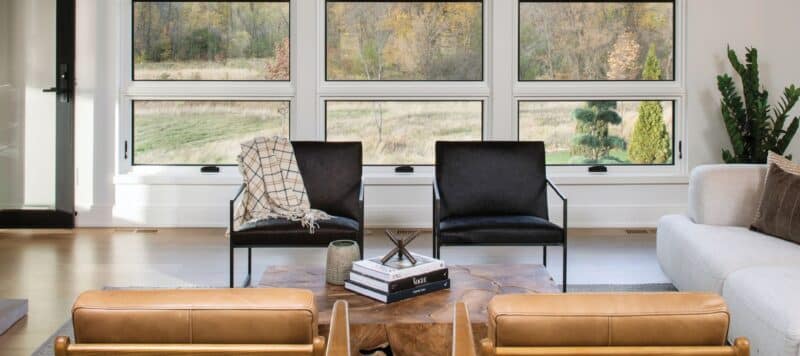 Modern living room with large windows showcasing a scenic outdoor view. The room features a seating area with black chairs, leather armchairs, and a wooden coffee table adorned with books and decorative items. A cozy blanket is draped over one of the chairs, adding to the inviting atmosphere
